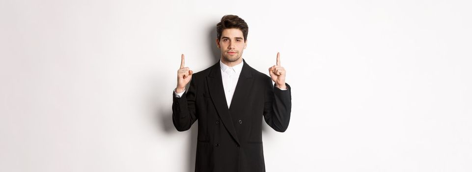 Image of confident and handsome man in formal suit, pointing fingers up, showing copy space on white background.