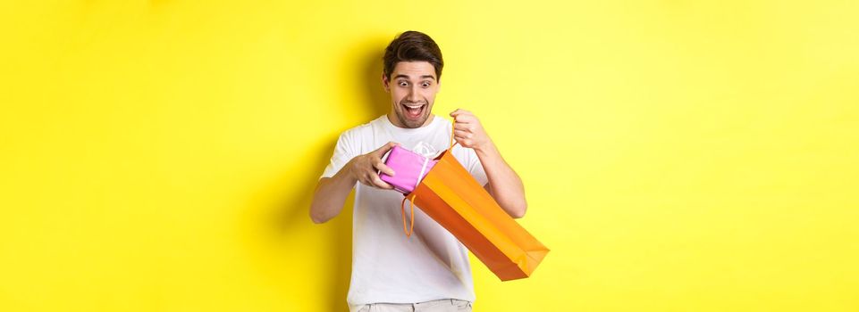 Concept of holidays and celebration. Young man looking surprised as take out gift from shopping bag, standing over yellow background.