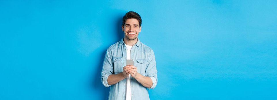 Handsome bearded man in casual outfit smiling at camera, checking smartphone, standing against blue background.