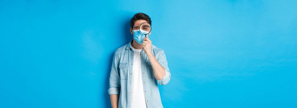 Concept of covid-19, social distancing and quarantine. Young man in medical mask searching for something, looking through magnifying glass, standing against blue background.