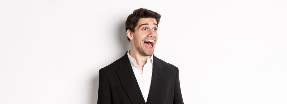 Close-up of handsome male entrepreneur in suit, looking left and smiling amazed, standing against white background.