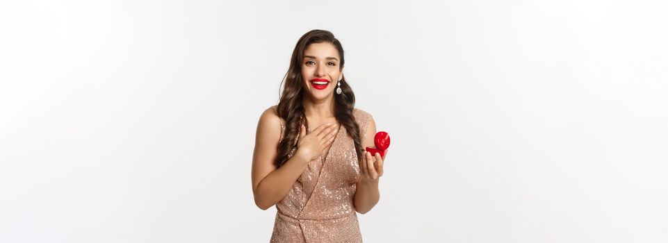 Image of beautiful woman in glamour dress receiving engagement ring, looking surprised and happy at camera, being proposed, standing over white background.