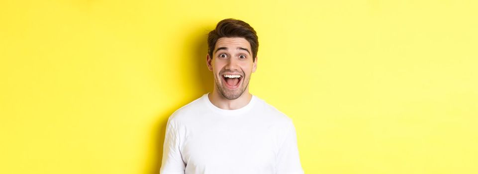 Close-up of surprised handsome guy reacting to great news, standing over yellow background in white t-shirt.