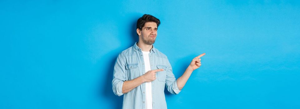 Doubtful adult man pointing fingers left at promotion and looking unsure, grimacing disappointed, standing against blue background.