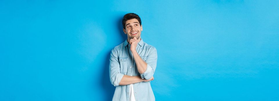 Portrait of thoughtful handsome man with beard, standing in casual outfit, looking at upper left corner and smiling, imaging or dreaming about something, standing over blue background.