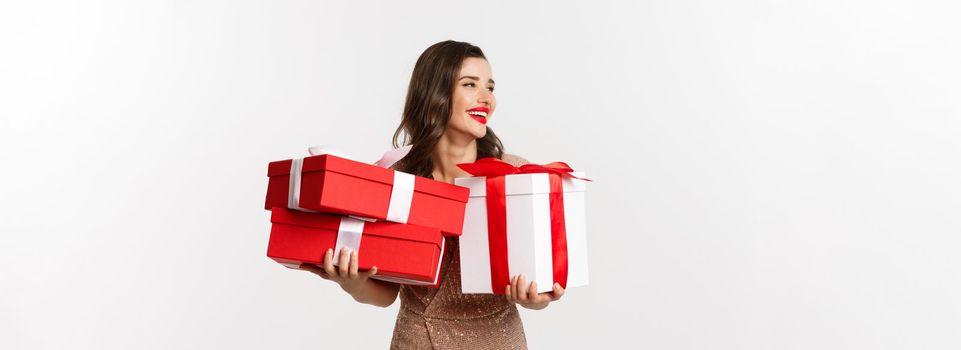 Holidays, celebration concept. Beautiful caucasian woman in elegant dress holding Christmas presents and smiling happy, standing over white background.