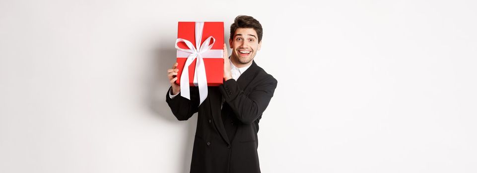 Handsome man in black suit, receiving christmas gift, smiling amazed, standing against white background.