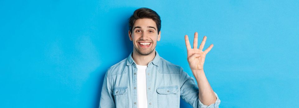 Close-up of handsome man smiling, showing fingers number four, standing over blue background.