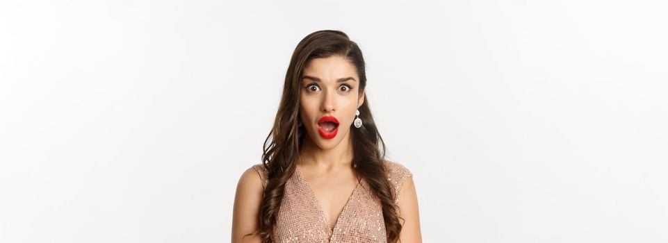 Image of attractive woman with red lips and hairstyle, wearing dress, staring surprised at camera, standing over white background.