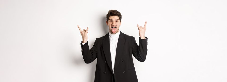 Portrait of happy attractive guy having fun at party, wearing black suit, showing rock-n-roll sign and tongue, standing excited against white background.