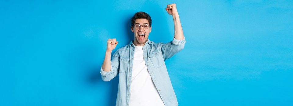 Excited handsome man triumphing, raising hands up and shouting for joy, celebrating victory, standing against blue background.