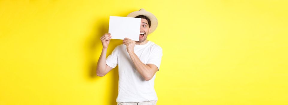 Excited man on vacation showing blank piece of paper for your logo, holding sign near face and smiling, standing against yellow background.