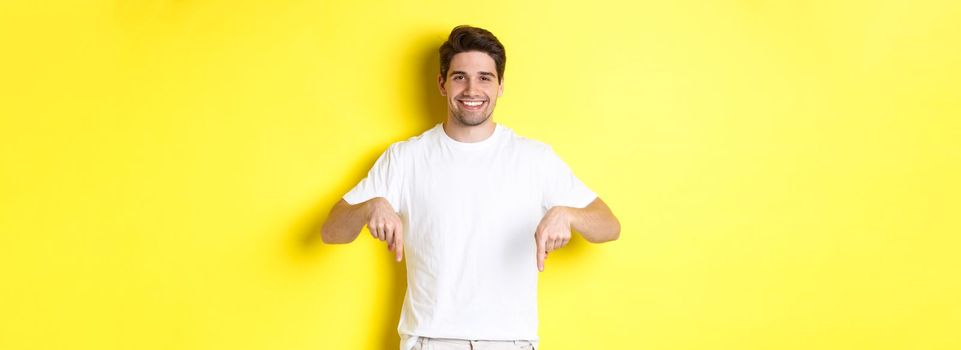 Attractive bearded man pointing fingers down, showing event banner, standing over yellow background.