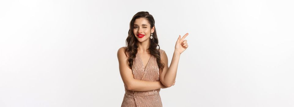 Christmas, holidays and celebration concept. Beautiful caucasian woman in party dress and red lipstick, winking and smiling, pointing finger left at promo offer, white background.