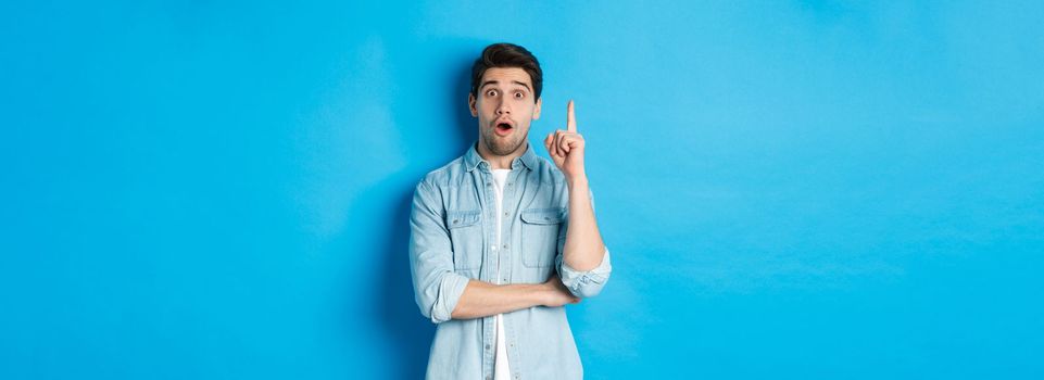 Image of handsome adult man having an idea, raising finger and smiling excited, found solution, standing against blue background.