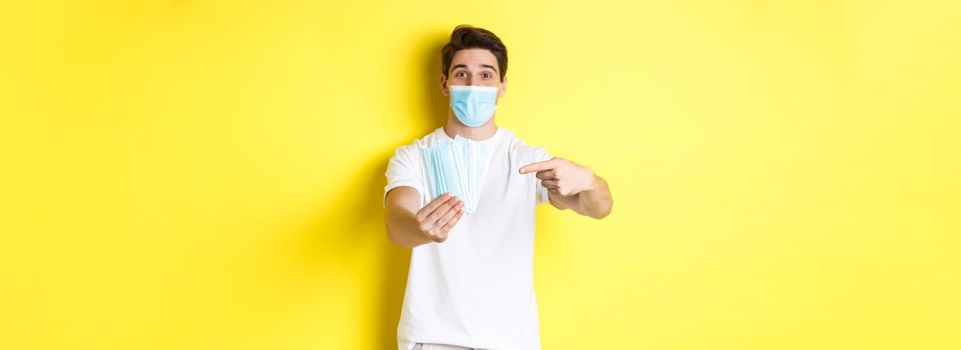 Concept of covid-19, quarantine and preventive measures. Young caucasian man giving medical masks for you, standing against yellow background.