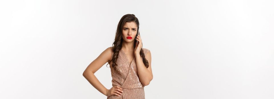 Christmas party and celebration concept. Elegant woman in glamour dress talking on phone and looking displeased, receive bad news, white background.
