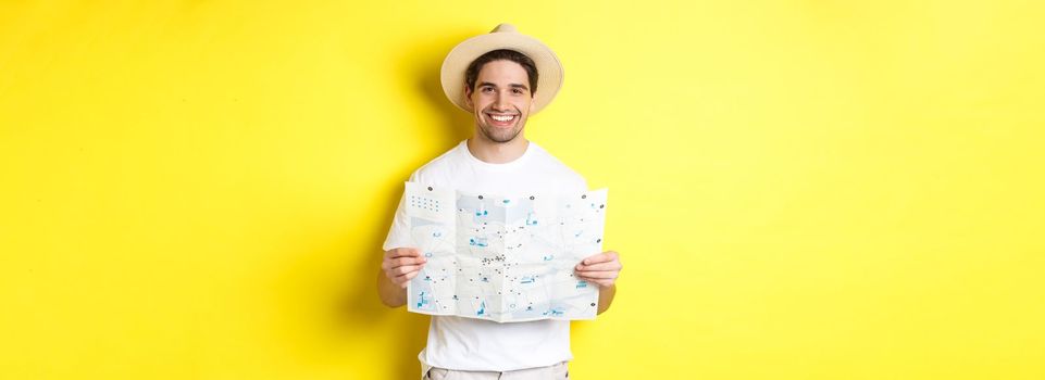 Travelling, vacation and tourism concept. Smiling young man going on trip, holding road map and smiling, standing over yellow background.