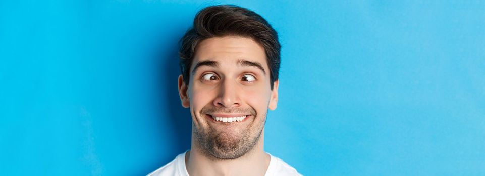 Head shot of young man making funny expressions, smiling and squinting, standing over blue background.