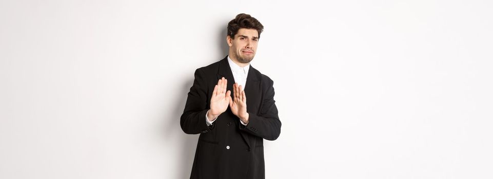 Image of stylish handsome man in black suit, cringe from something embarrassing, declining offer and showing stop gesture, standing against white background.