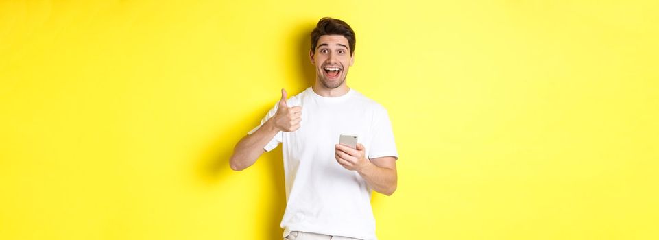 Happy smiling man holding smartphone, showing thumb up in approval, recommend something online, standing over yellow background.