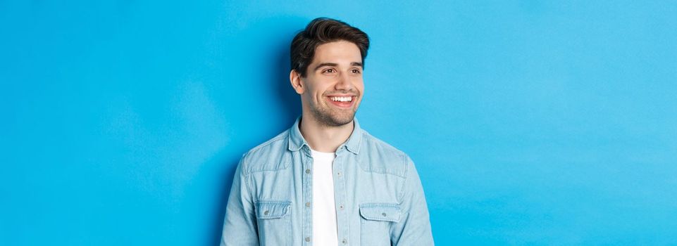 Young modern guy with beard, looking left and smiling pleased at copy space, standing over blue background.