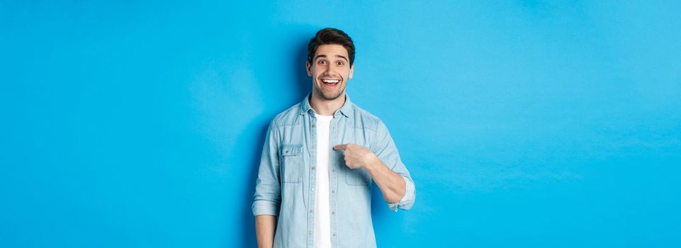 Happy and surprised man pointing at himself, smiling pleased, standing against blue background.