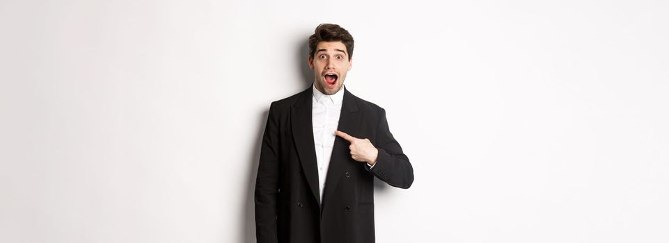 Concept of new year party, celebration and lifestyle. Portrait of surprised handsome guy in black suit, pointing at himself and looking amazed, being chosen, standing over white background.