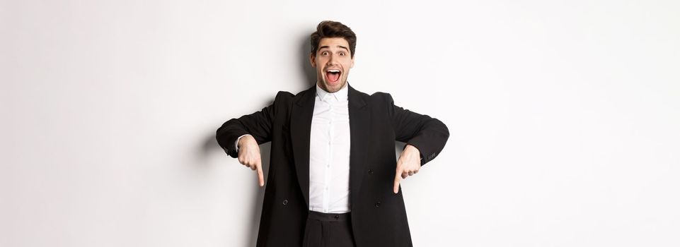 Portrait of super happy man showing christmas promo offer, pointing fingers down and smiling amazed, wearing formal party suit, standing over white background.