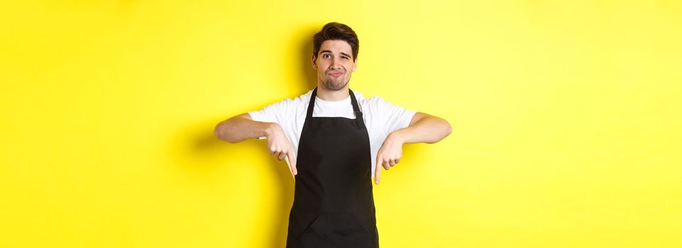 Skeptical barista in black apron pointing fingers down at bad product, looking displeased and unamused, standing over yellow background.