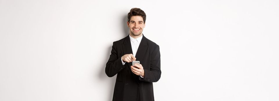 Image of handsome businessman in suit showing you app on smartphone, pointing finger at mobile screen and smiling, standing over white background.