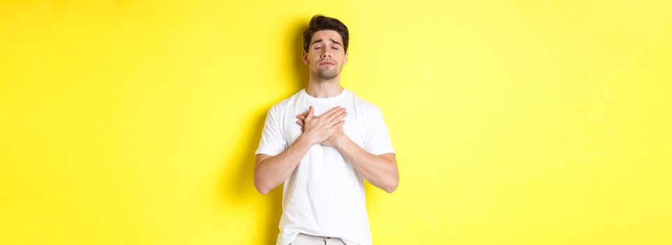 Romantic and nostalgic man holding hands on heart, close eyes and remember something, standing over yellow background.