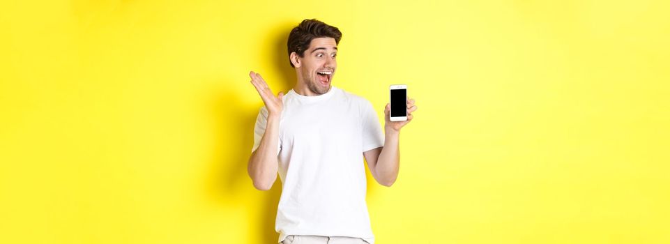 Image of amazed guy looking at mobile phone screen with surprised face, standing excited against yellow background.