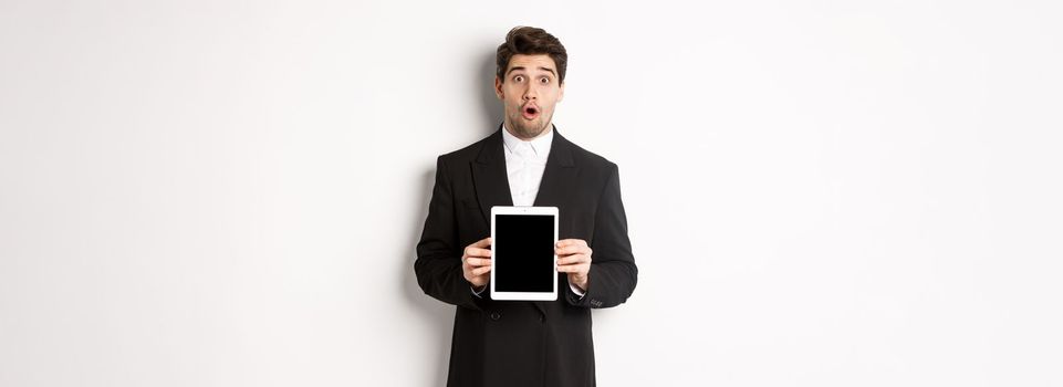 Image of surprised handsome man in black suit, showing digital tablet screen and looking amazed, standing against white background.