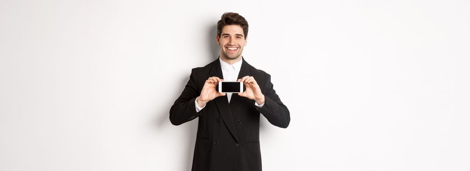 Portrait of attractive businessman in black suit, holding smartphone horizontally and showing screen, smiling pleased, standing against white background.