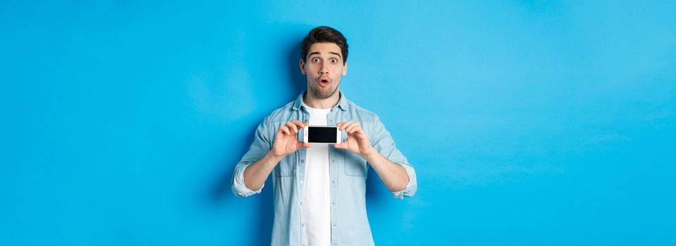 Surprised guy showing mobile phone screen and looking impressed, standing over blue background.