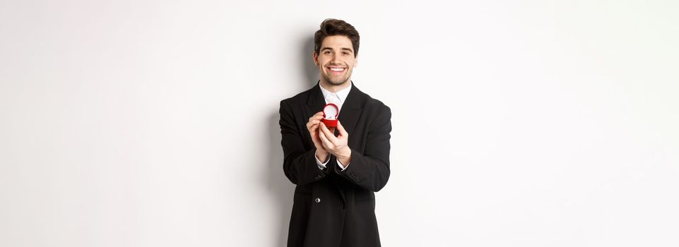 Image of handsome boyfriend in black suit making a proposal, asking to marry him and showing wedding ring, standing over white background.