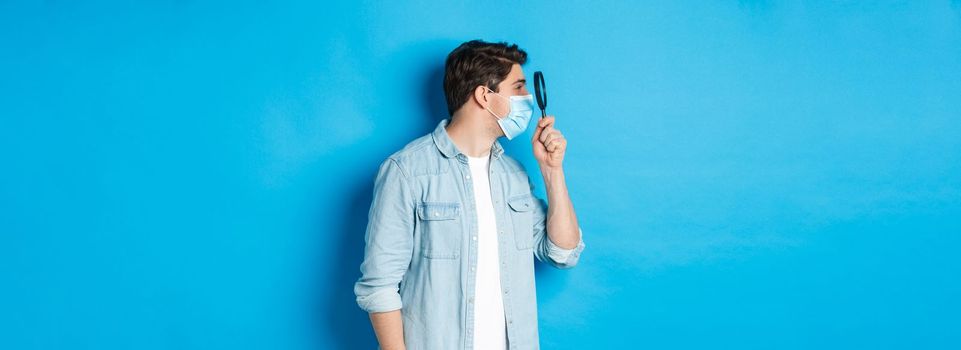 Concept of covid-19, social distancing and quarantine. Young man in medical mask searching for something, looking left through magnifying glass, standing against blue background.