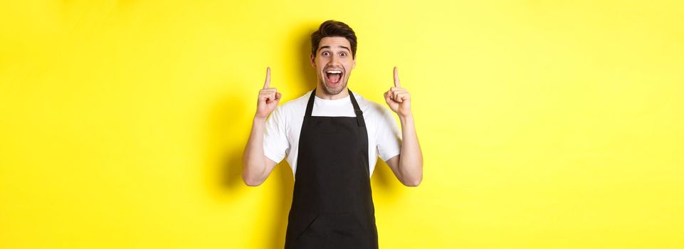 Excited coffee shop owner in black apron pointing fingers up, showing special offers, standing over yellow background.