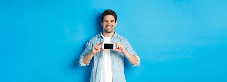 Handsome smiling man showing smartphone screen, standing against blue background for copy space.