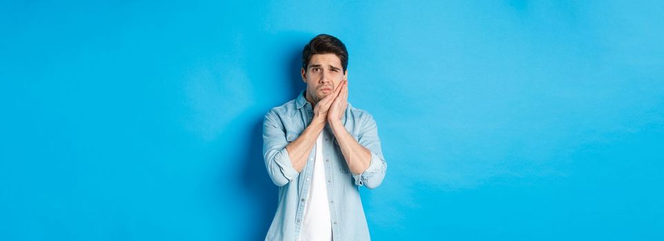 Portrait of man with toothache grimacing from pain and touching cheek, standing against blue background.