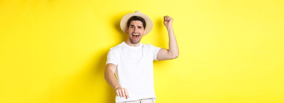 Concept of tourism and summer. Young man traveller showing rodeo gesture, standing in straw hat and white clothes, standing over yellow background.