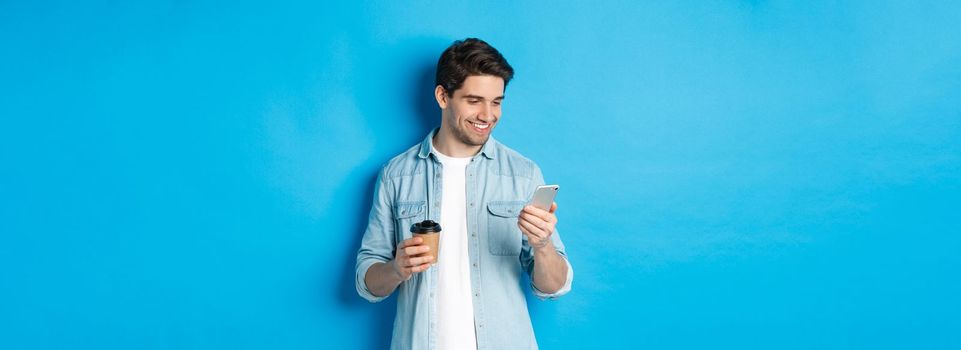 Handsome modern guy reading message on cell phone and drinking coffee, standing against blue background.