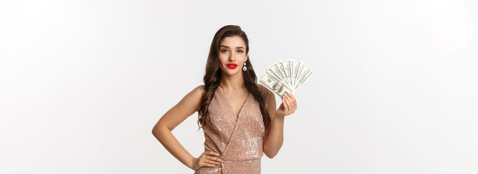 Shopping concept. Sassy beautiful woman with red lipstick, wearing glamour dress, showing money dollars and looking confident, standing over white background.