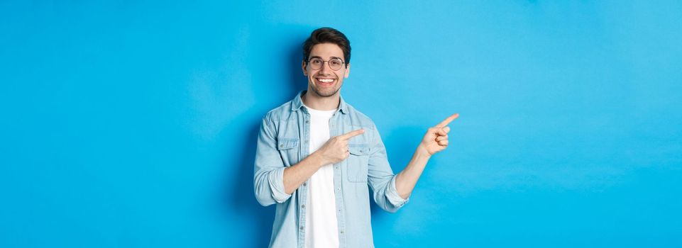 Young handsome man in glasses showing advertisement, smiling and pointing fingers left, making announcement, standing against blue background.