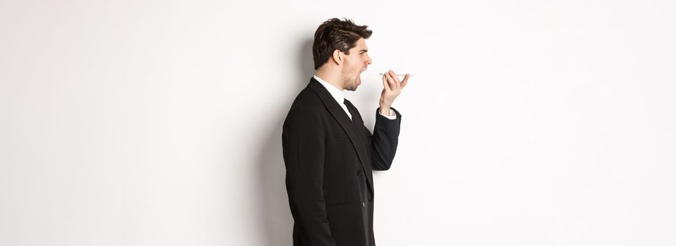 Profile shot of angry businessman in black suit, shouting at speakerphone and looking mad, recording voice message, standing over white background.