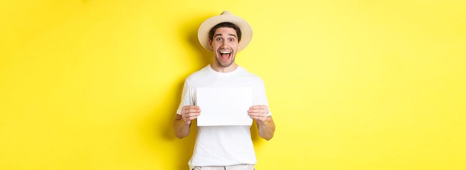 Excited tourist showing your logo or sign on blank piece of paper, smiling amazed, standing against yellow background.