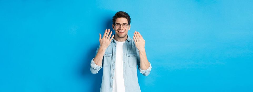 Handsome young man in glasses showing manicure hands and smiling, taking care of nails, standing over blue background.