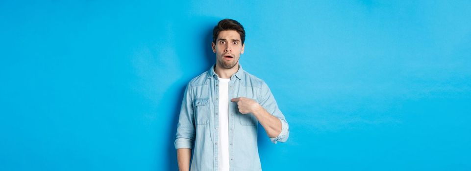 Nervous man pointing at himself and looking confused, standing in casual clothes over blue background.