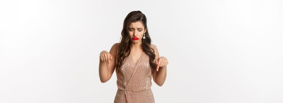 Sad beautiful girl pointing fingers down and sulking, wearing dress for christmas party, looking disappointed, standing over white background.